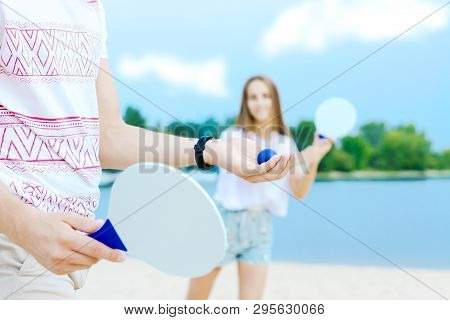 Male Is Holding Blue Ball, His Smiling Girlfriend Ready To Catch With Racket On Background. Young Ma