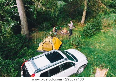 Couple Sitting Near Campfire Drinking Tea. Yellow Tent And White Suv Car. Active Leisure Time