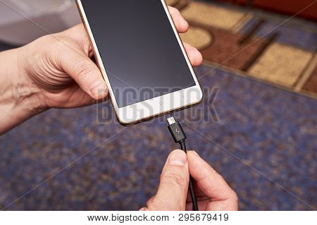 Female Hand Inserts The Usb Cable Charger Into The Mobile Phone Against The Background Of The Apartm