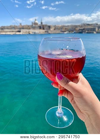 A Glass Of Cherry Drink, Valletta Panoramic View And Bright Turquoise Meditarranean Sea Background, 