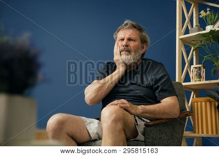 Inflammation. Old Bearded Man Sitting On The Chair In Black Shirt And Suffering From Teeth Pain On B