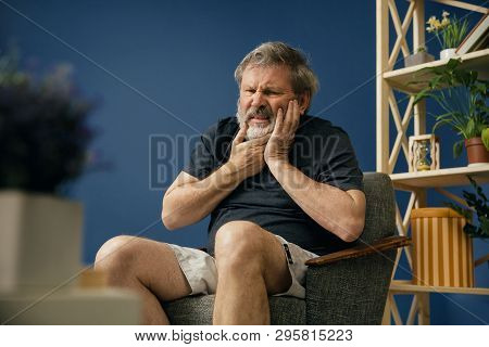 Loosing His Face And Feelings. Old Bearded Man In Black Shirt Sitting On The Chair And Suffering Fro
