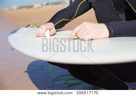 Closeup Of Young Man Waxing Surfboard On Sunny Beach. Guy Wearing Wetsuit, Crouching And Holding Sur