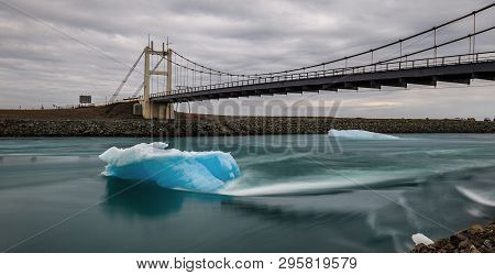 An Ice Berg Lying In A River Under A Bridge In Iceland.