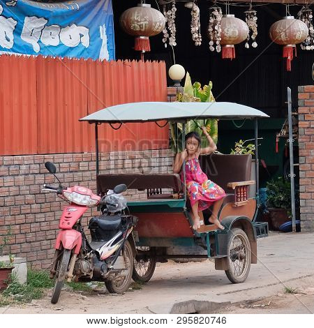 Cambodia, Siem Reap 12/08/2018 A Little Asian Girl Sits In A Moto Rickshaw Near A House With Red Lan