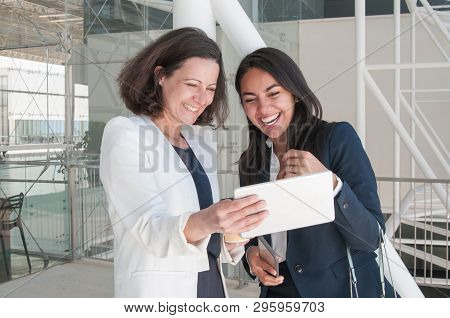 Two Smiling Business Women Using Tablet In Office Hall. Colleagues Or Partners Standing And Videocon