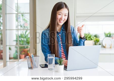 Beautiful Asian woman working using computer laptop happy with big smile doing ok sign, thumb up with fingers, excellent sign