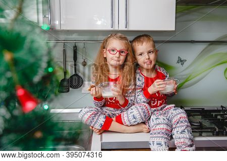Little Brother And Sister Sit On A Christmas-decorated Kitchen Table In Pajamas And Drink Milk. Girl