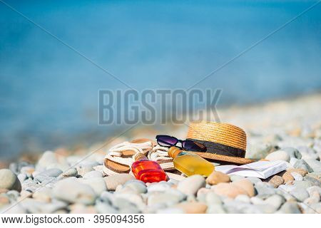 Beach Hat On Opened Book With Sunscreen And Shoes On Pebble Beach