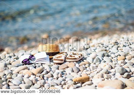 Beach Hat On Opened Book With Sunscreen And Shoes On Pebble Beach