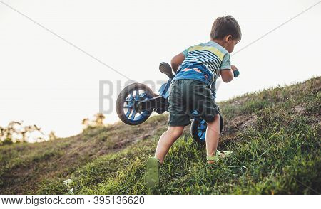 Caucasian Boy Is Carrying The Bike On A Hill While Playing In A Field During A Summer Evening