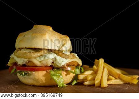 Close Up Of A Homemade Burger With Onion Rings On A Chopping Wood Board, On Black Background