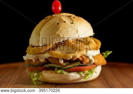 Close Up Of A Homemade Chicken Burger With Onion Rings On A Chopping Wood Board, On Black Background