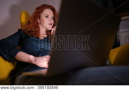 Nice beautiful lady with redhead curly hair work at the notebook sit down on the sofa at home - check on oline shops for cyber monday sales - technology woman concept for alternative office freelance