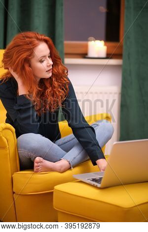 Nice beautiful lady with redhead curly hair work at the notebook sit down on the sofa at home - check on oline shops for cyber monday sales - technology woman concept for alternative office freelance