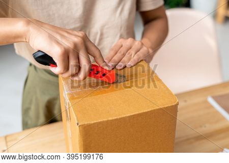 Hands of young female with stationery knife cutting sealed cellotape on top of packed cardboard box while standing by table during work
