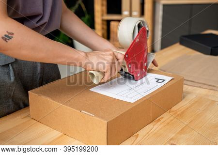 Hands of young female manager of online shop with cellotape gun dispenser sealing cardboard box with packed order of client by table