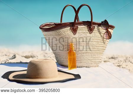 Women's beach accessories on sand for summer vacation concept. Straw tote bag, sun hat and sunscreen lotion or suntan tanning oil spray bottle with blue ocean background for travel holidays.