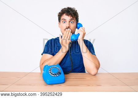 Young caucasian man with curly hair using vintage telephone sitting on the table covering mouth with hand, shocked and afraid for mistake. surprised expression 