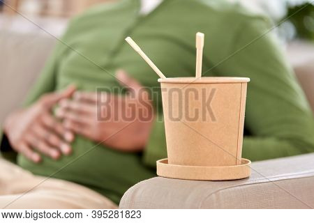 consumption and people concept - cup of takeaway food in front of overeaten man at home