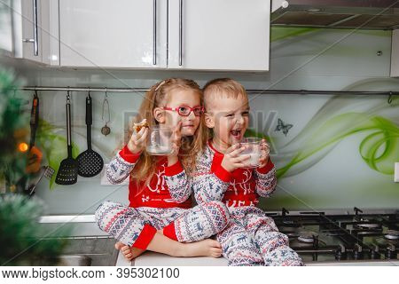 Little Brother And Sister Sit On A Christmas-decorated Kitchen Table In Pajamas And Drink Milk. Girl