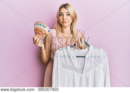 Young caucasian woman holding hanger with t shirt and euro banknotes in shock face, looking skeptical and sarcastic, surprised with open mouth 