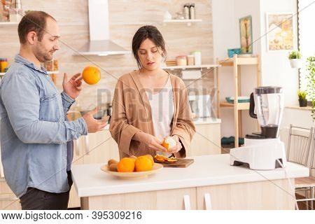 Couple Preparing Fresh Smoothie In Kitchen. Woman Peeling Off Oranges. Husband Doing Tricks With Fru