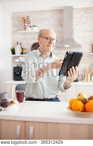 Happy Old Man In Kitchen During Breakfat Using Tablet Pc. Elderly Person With Tablet Portable Pad Pc