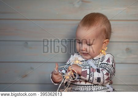 Baby Girl Wearing Clothes With Christmas Print Playing With Lights On Light Wooden Background. New Y