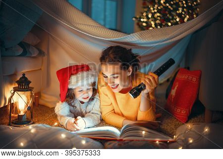 Merry Christmas! Pretty young mother reading a book to her daughter near Christmas tree. 