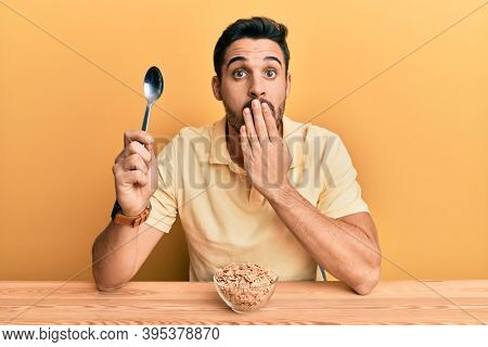 Young hispanic man eating healthy whole grain celears sitting on the table covering mouth with hand, shocked and afraid for mistake. surprised expression 