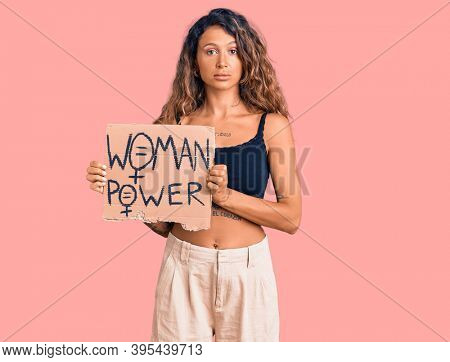 Young hispanic woman with tattoo holding woman power banner thinking attitude and sober expression looking self confident 