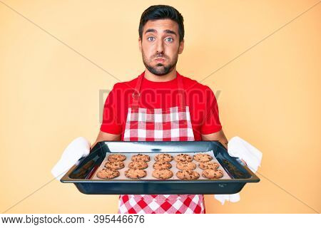 Young hispanic man wearing baker uniform holding homemade cookies puffing cheeks with funny face. mouth inflated with air, catching air. 
