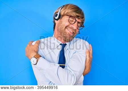 Young blond man wearing call center agent headset hugging oneself happy and positive, smiling confident. self love and self care 