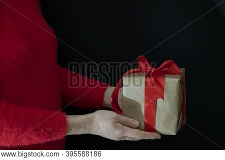 Female Hands In A Red Sweater Hold A Gift Wrapped In Craft Paper With A Wide Red Ribbon On A Black B