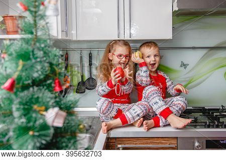 Little Laughing Twins Sit On The Table In Pajamas In The Christmas Decorated Kitchen With An Apple. 