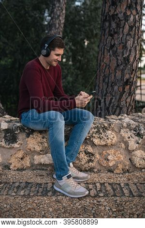 Boy Listening To Music With Wireless Headphones And Using His Cell Phone. Red Vest.