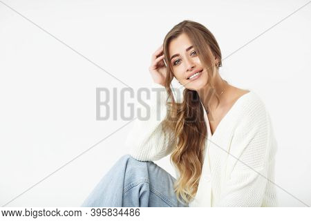 Beautiful Young Woman In Blue Jeans And White Sweater Sitting On A White Background. Portrait Of A C