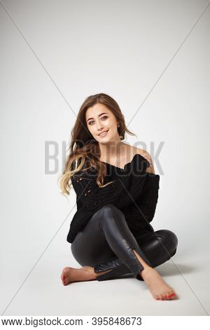 Beautiful Young Woman In Black Leather Leggings And Black Sweater Sitting On A White Background. A C