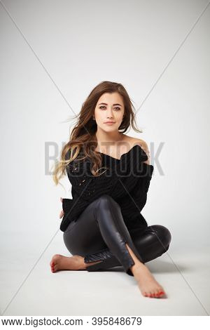 Beautiful Young Woman In Black Leather Leggings And Black Sweater Sitting On A White Background. A C
