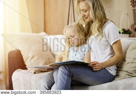 Happy Family. Blonde Young Mother Reading A Book To Her Cute Daughter While Sitting At Wooden Floor 