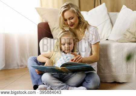 Happy Family. Blonde Young Mother Reading A Book To Her Cute Daughter While Sitting At Wooden Floor 