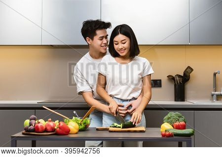 Loving Chinese Husband Hugging Wife Cooking Dinner In Modern Kitchen At Home, Making Veggetable Sala