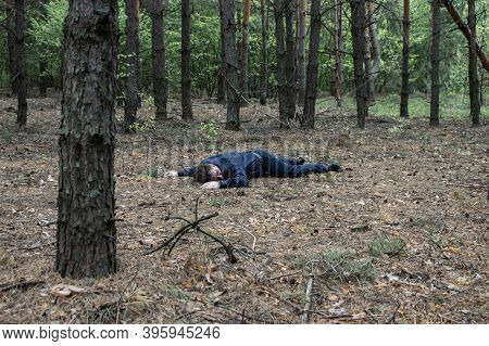 Murder In The Woods. The Body Of A Man In A Blue Shirt And Trousers Lies On The Ground Among The Tre