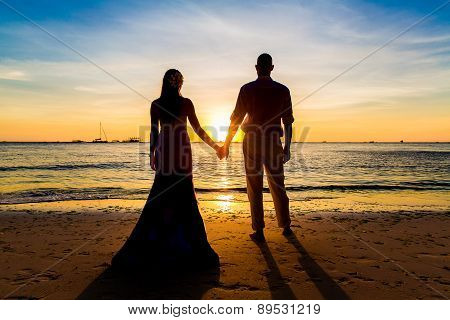 The Bride And Groom Are Kept Hands On A Tropical Beach . Silhouette Photo At Sunset .