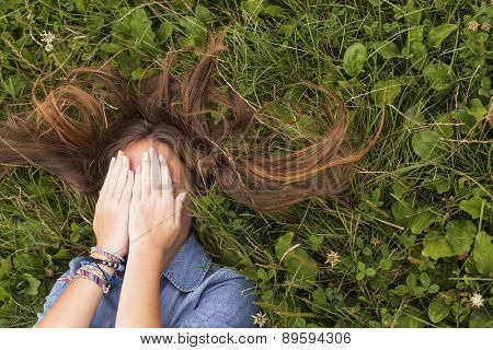Young cute girl with tousled long hair lying on the green grass covering his face with his hands.