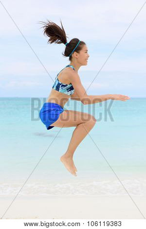 Crossfit fitness Asian woman on beach doing jump squat plyometric training exercises by jumping and touching knees as part of a full body core workout and active lifestyle.