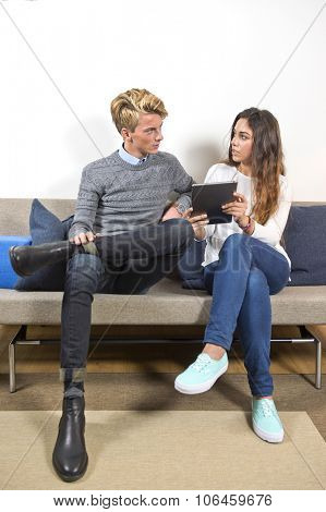 Young man and woamn, sitting on a couch in a living room, surfing together on a tablet. The woman is showing him something online.