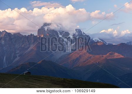 Mountain landscape. The top of Ushba in clouds. Beautiful sunset. Wooden house of shepherds on a hillside Mkheer. Caucasus, Zemo Svaneti, Georgia 