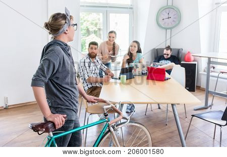 Active young woman holding a commuter bike while talking with her colleagues in the modern office of a shared co-working space for independent workers